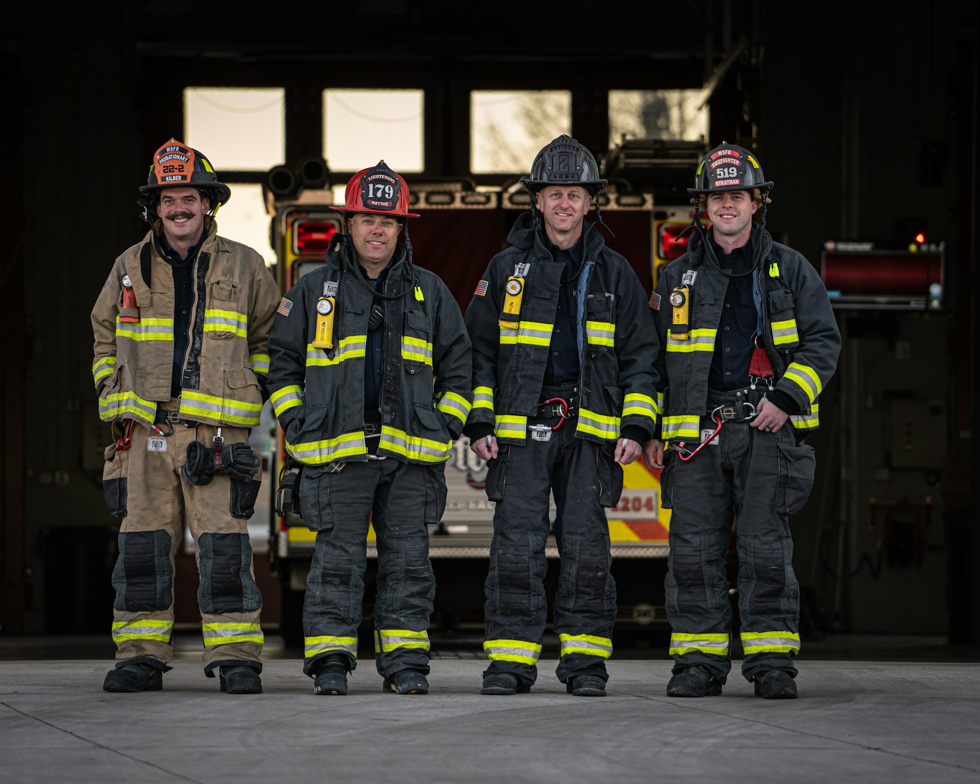 Four firefighters in gear standing in front of a fire truck.