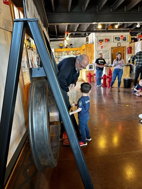 An adult and a child stand in front of a hanging train tire, which is the tool used by firefighters before telephones to signal an emergency.