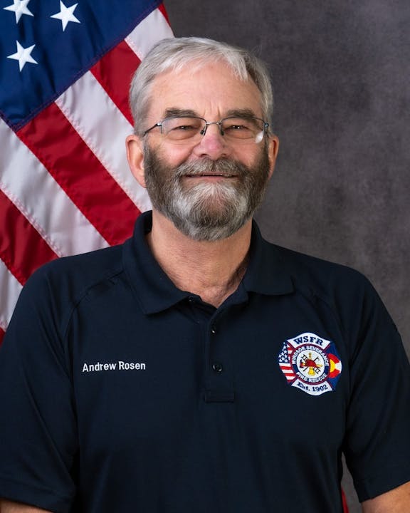 A man with glasses in a polo shirt with a badge, standing before an American flag.