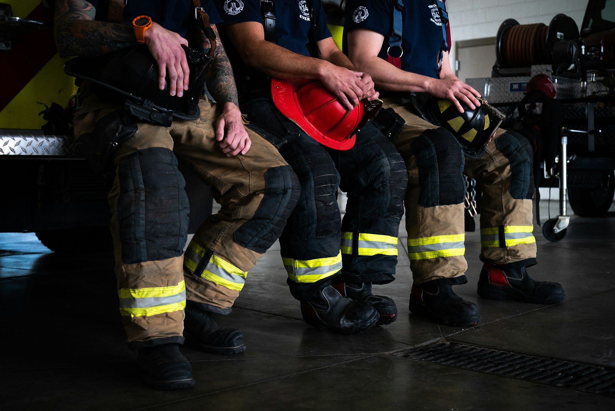 Firefighters in gear sitting, with helmets on laps, in front of a fire truck.