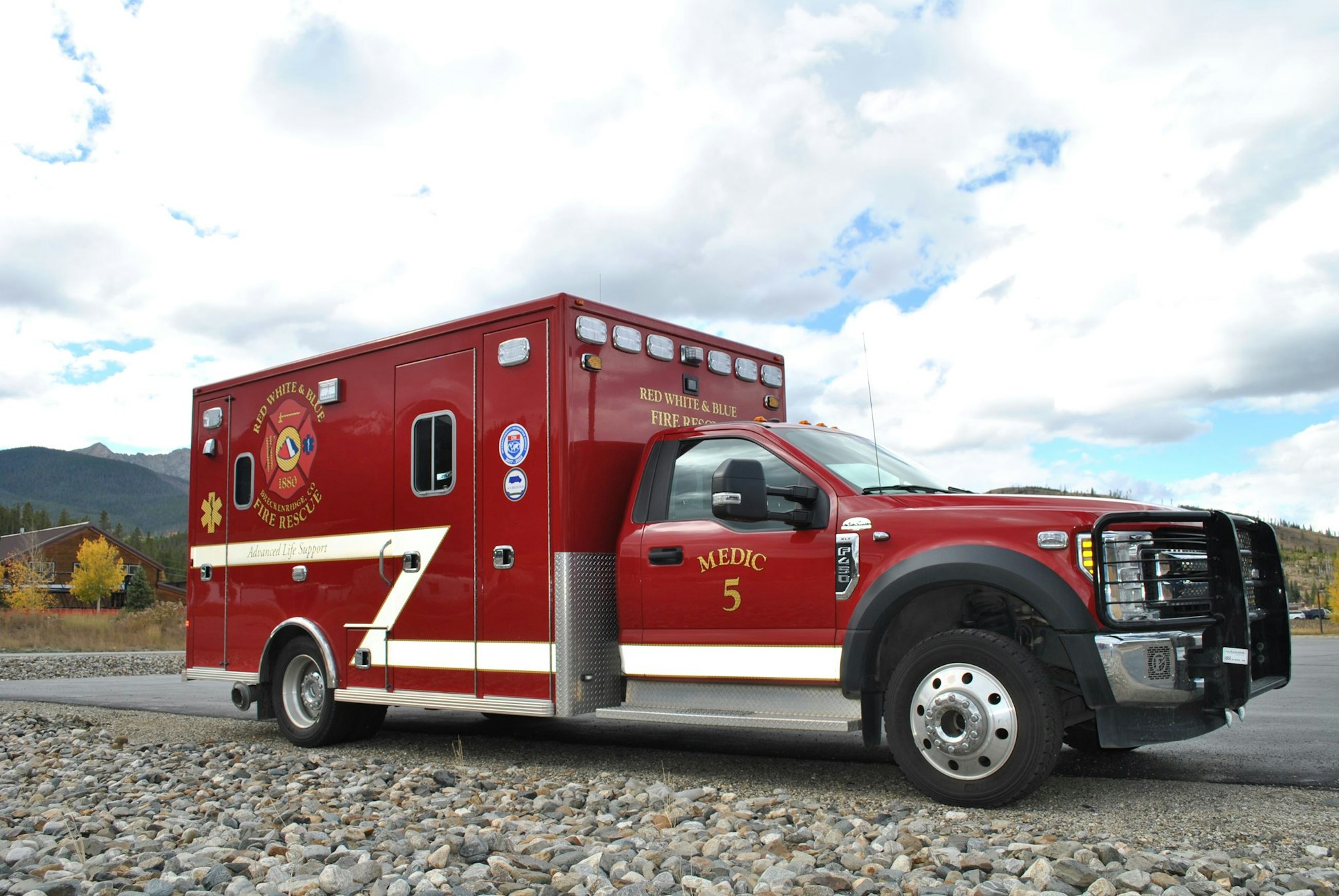 A red ambulance with "Red White & Blue Fire Rescue" text, parked on gravel with mountains and cloudy sky in the background.