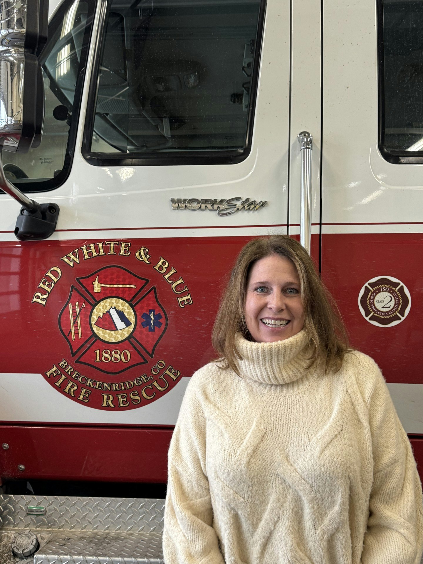 A person in a cream sweater stands in front of a fire truck labeled "Red, White & Blue Fire Rescue, Breckenridge, CO."