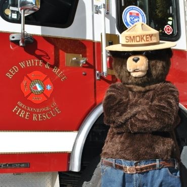 A person in a Smokey Bear costume stands in front of a fire truck.