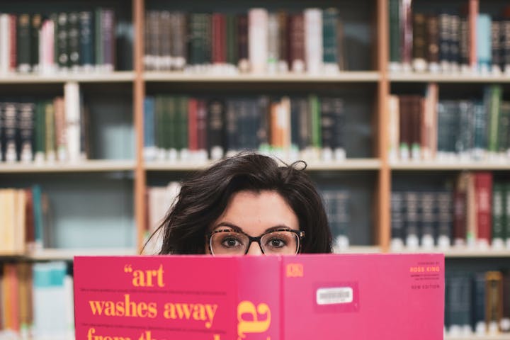 May contain: book, indoors, library, publication, shelf, person, reading, accessories, glasses, and furniture