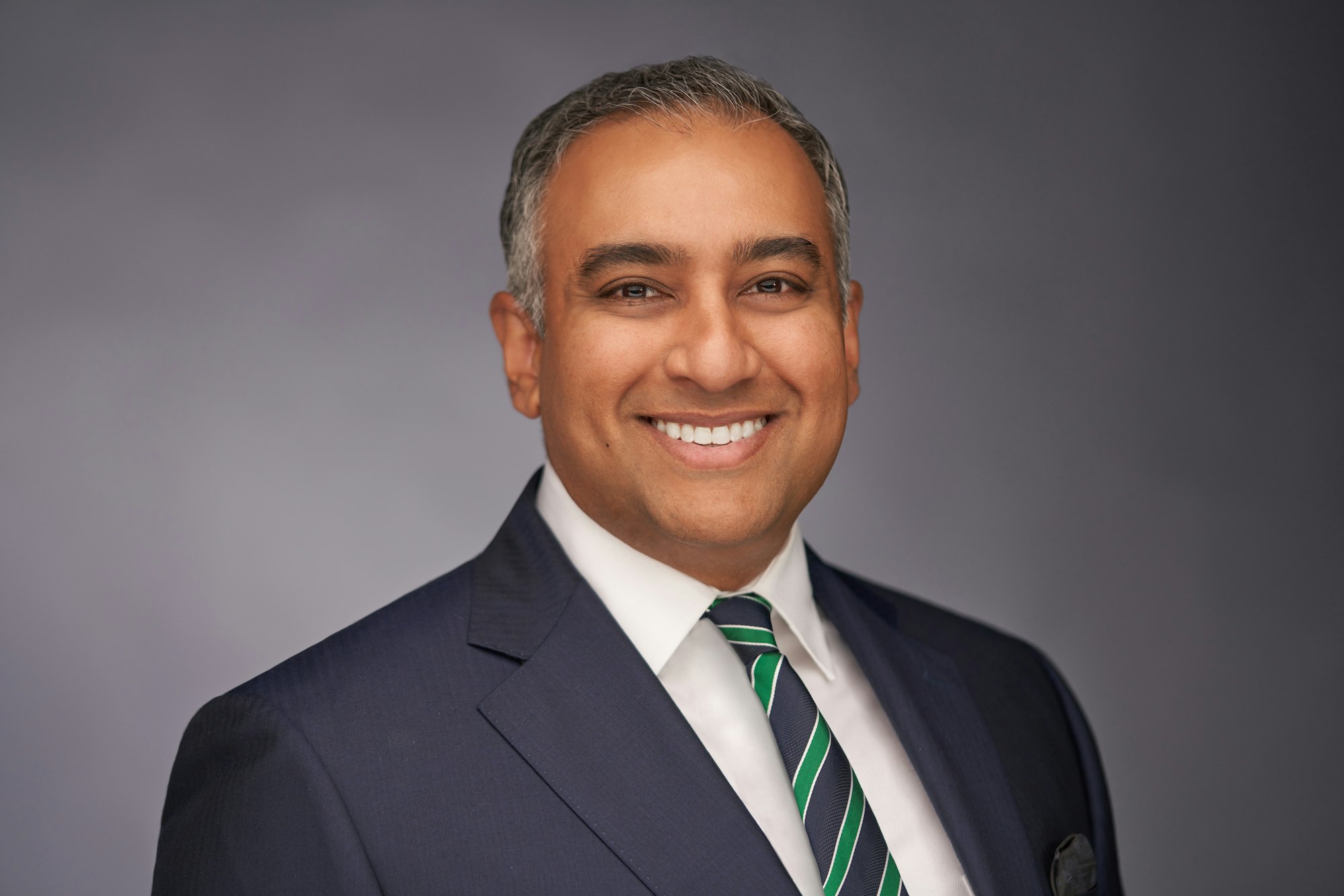 A smiling person in a suit with a striped tie, on a grey background.