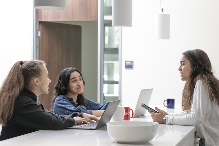 Three young women are engaged in a discussion at a table with laptops and mugs in a modern, bright setting.