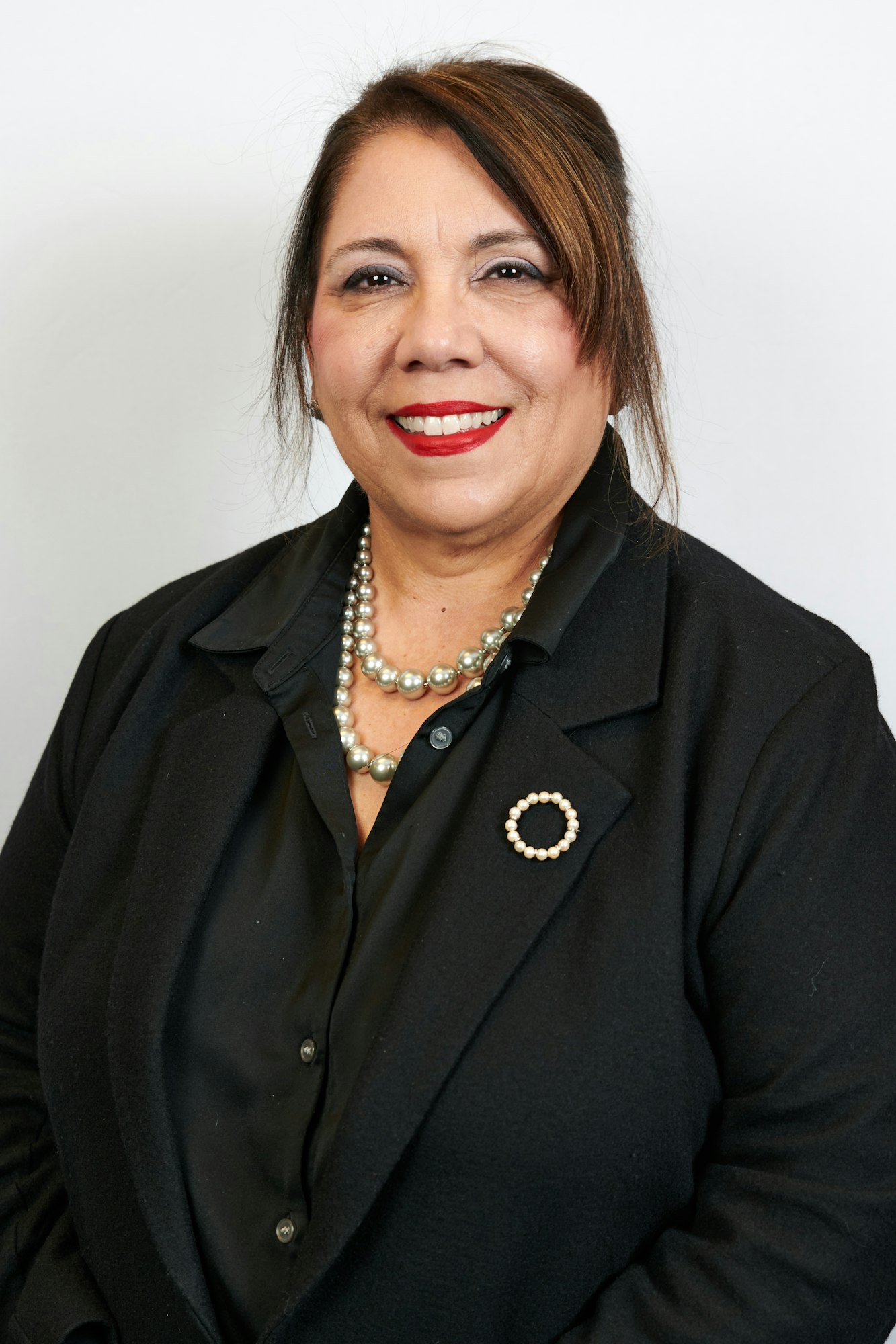 A smiling woman with red lipstick, wearing a black shirt, a pearl necklace, and a brooch.