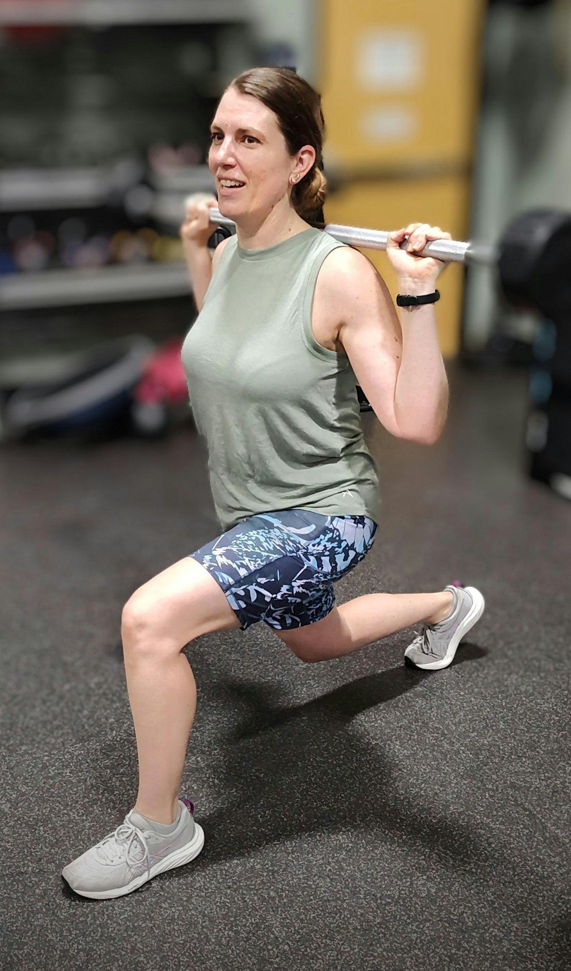 A person doing a barbell lunge exercise in a gym, wearing a tank top and patterned shorts.