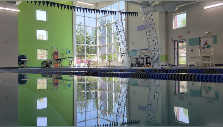 A bright indoor pool area with green walls, large windows, diving equipment, and reflection on the water's surface.