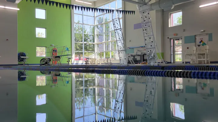 A bright indoor pool area with green walls, large windows, diving equipment, and reflection on the water's surface.