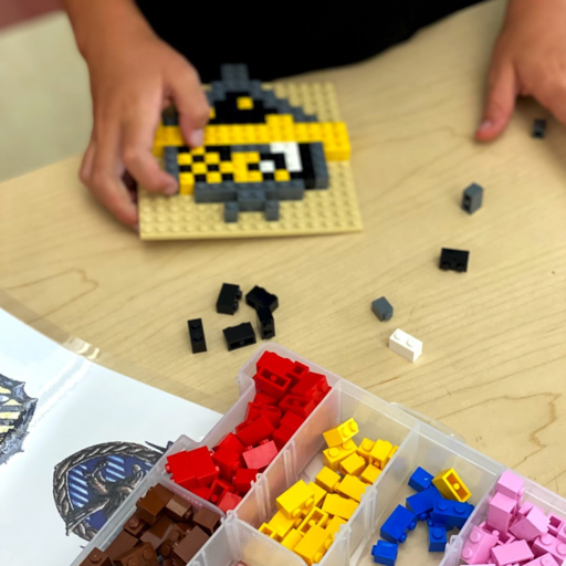 Hands assembling a pixel art creation with LEGO bricks on a tan baseplate, colorful LEGO pieces nearby.