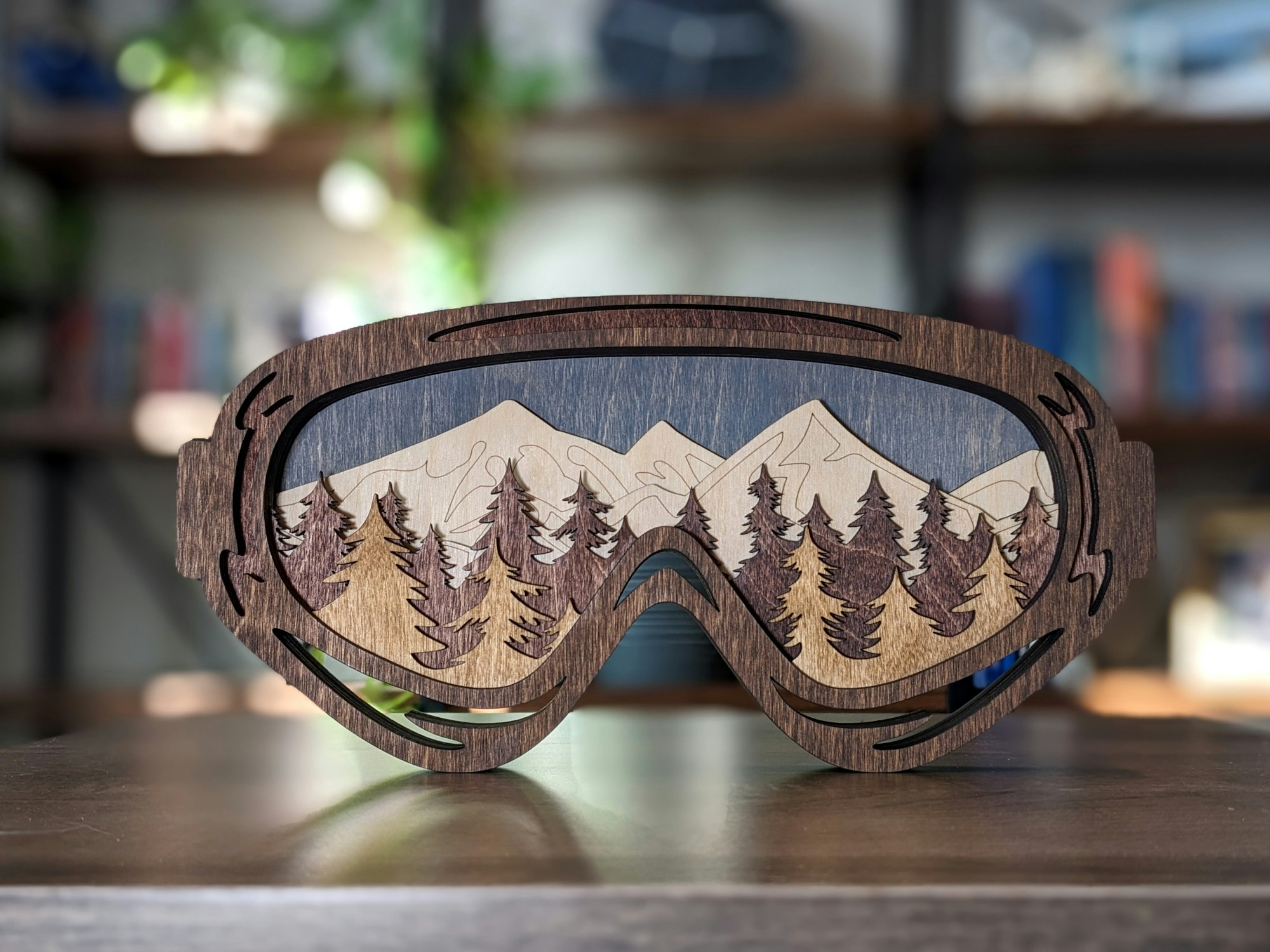 Wooden artwork shaped like goggles, depicting a mountain landscape with trees.