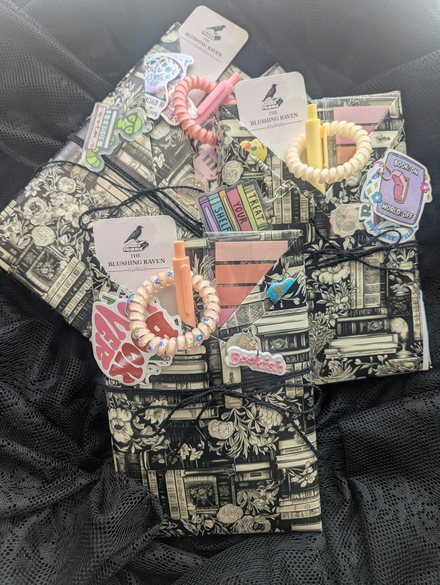 Notebooks with vintage book print covers, accessories, and stickers, including pens, coiled bracelets, and brand tags on black fabric.