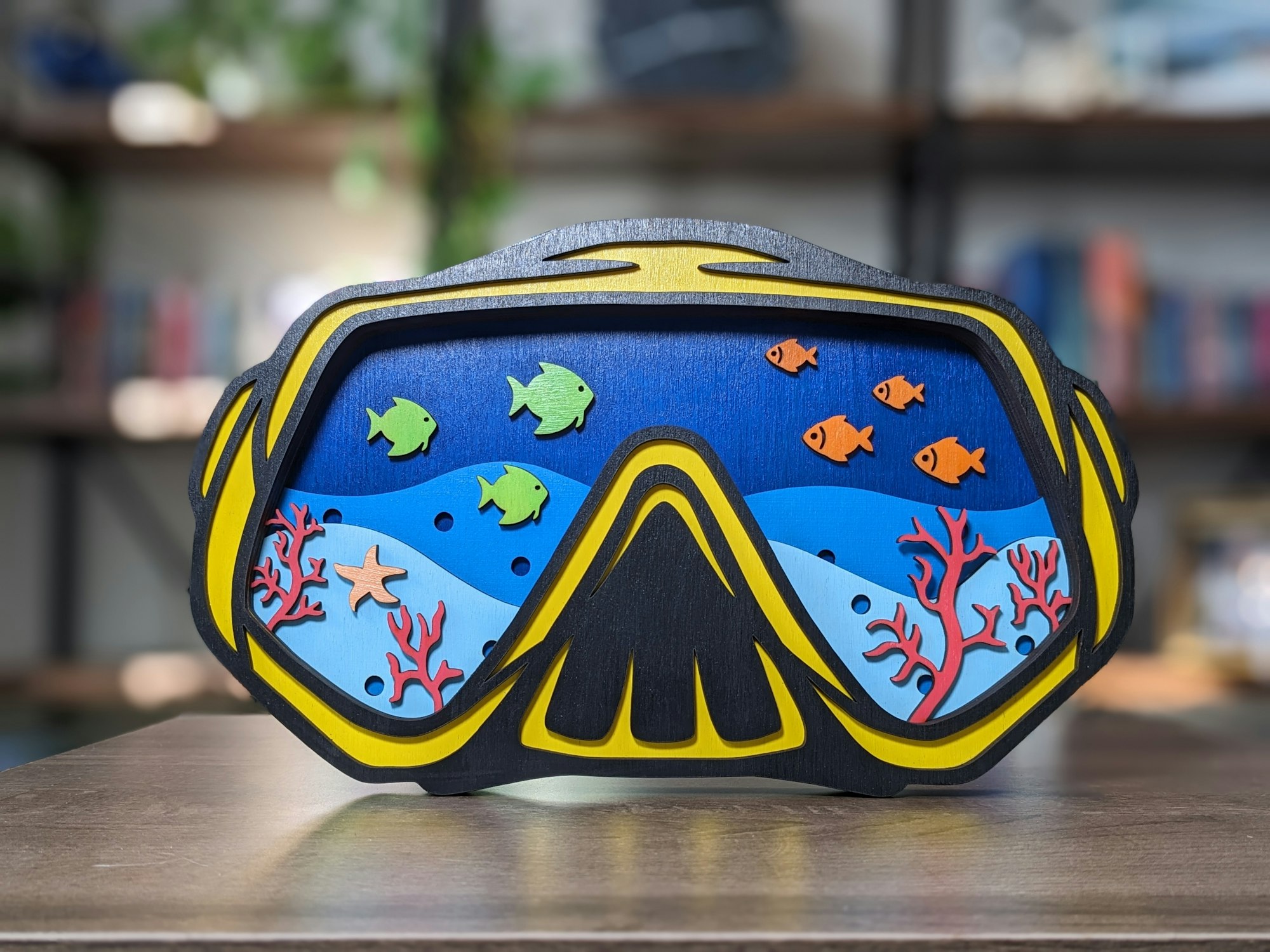 A decorative scuba mask with an ocean scene featuring colorful fish, corals, and a starfish on a wooden table.