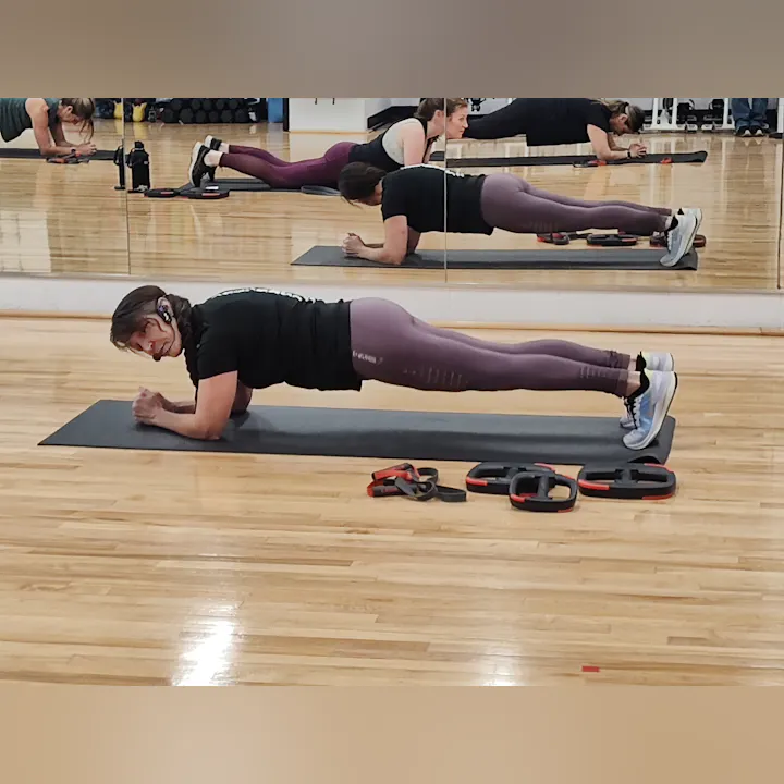 A person is performing a plank exercise on a mat in a gym, with workout equipment visible in the background.