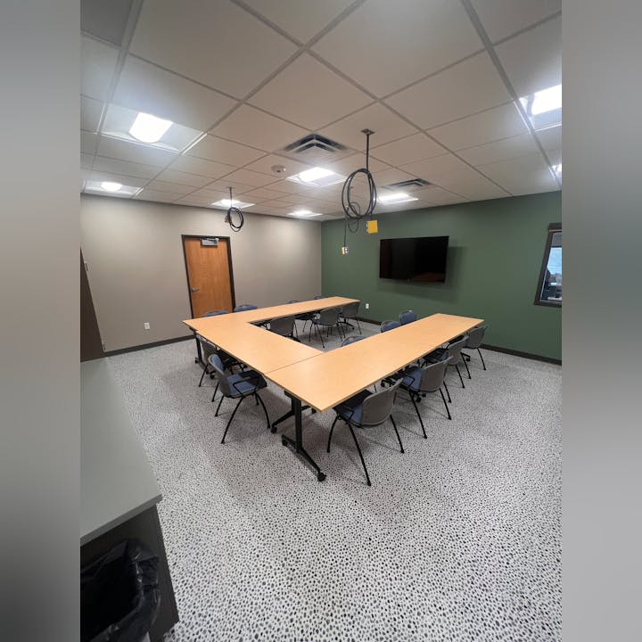 A conference room with a large table, chairs, a TV on the wall, and a speckled floor.