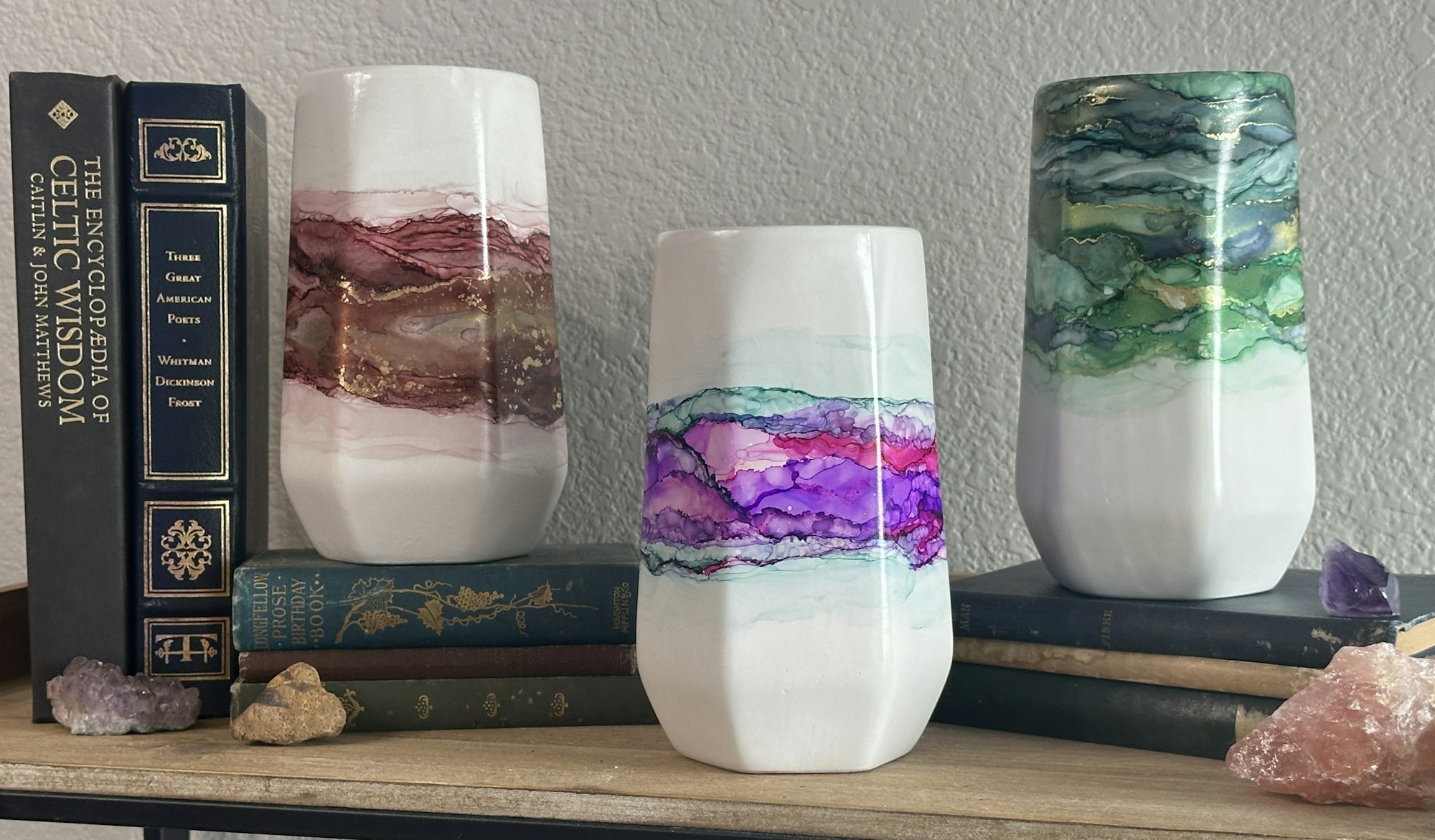 Three watercolor vases with abstract designs on a shelf, surrounded by books and crystals.