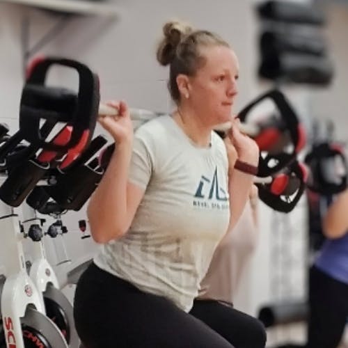 A woman is lifting weights in a gym, positioned near exercise bikes.