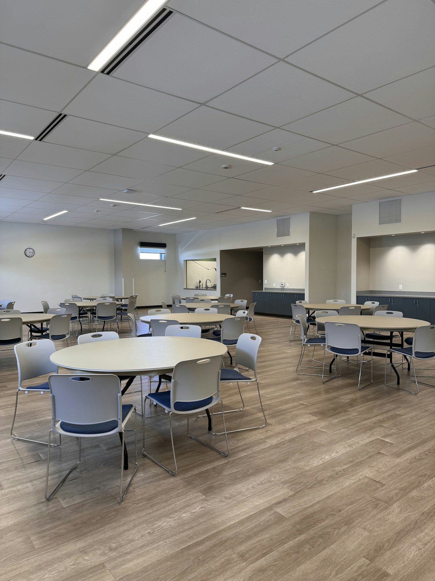 A room with round tables and chairs on a wooden floor, ceiling with lights and a clock.