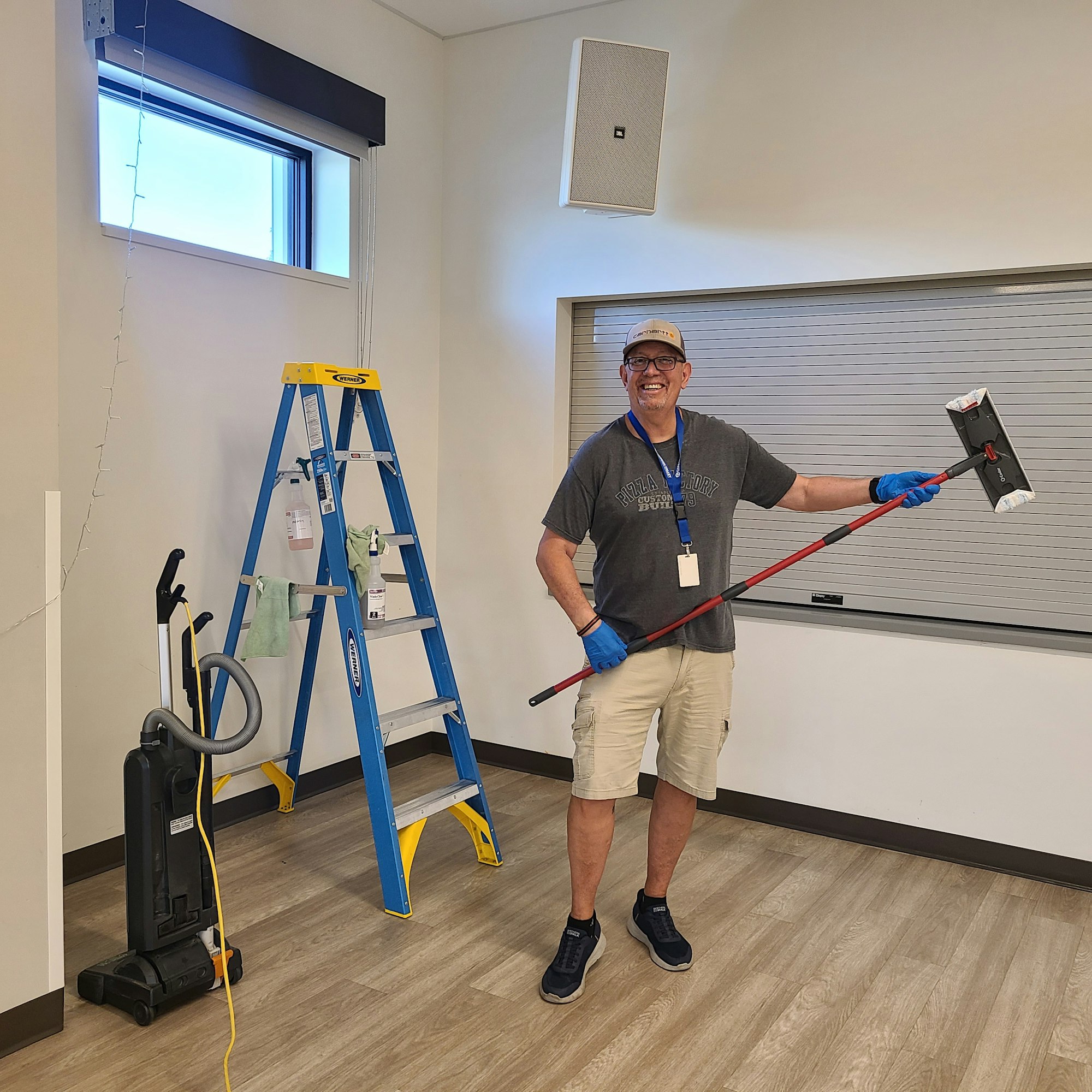 A smiling person in gloves holds a cleaning tool near a ladder and vacuum in a bright, empty room.