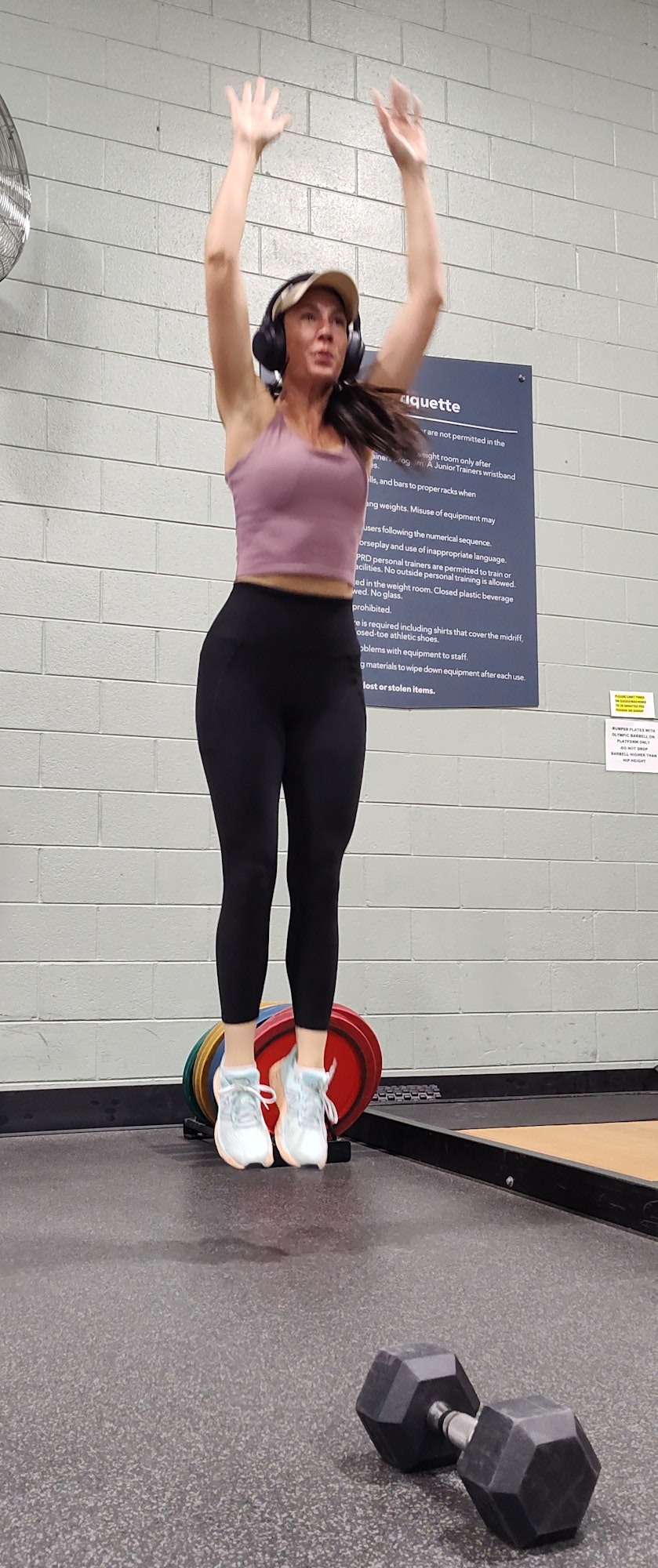 A woman is jumping in a gym with dumbbells nearby.