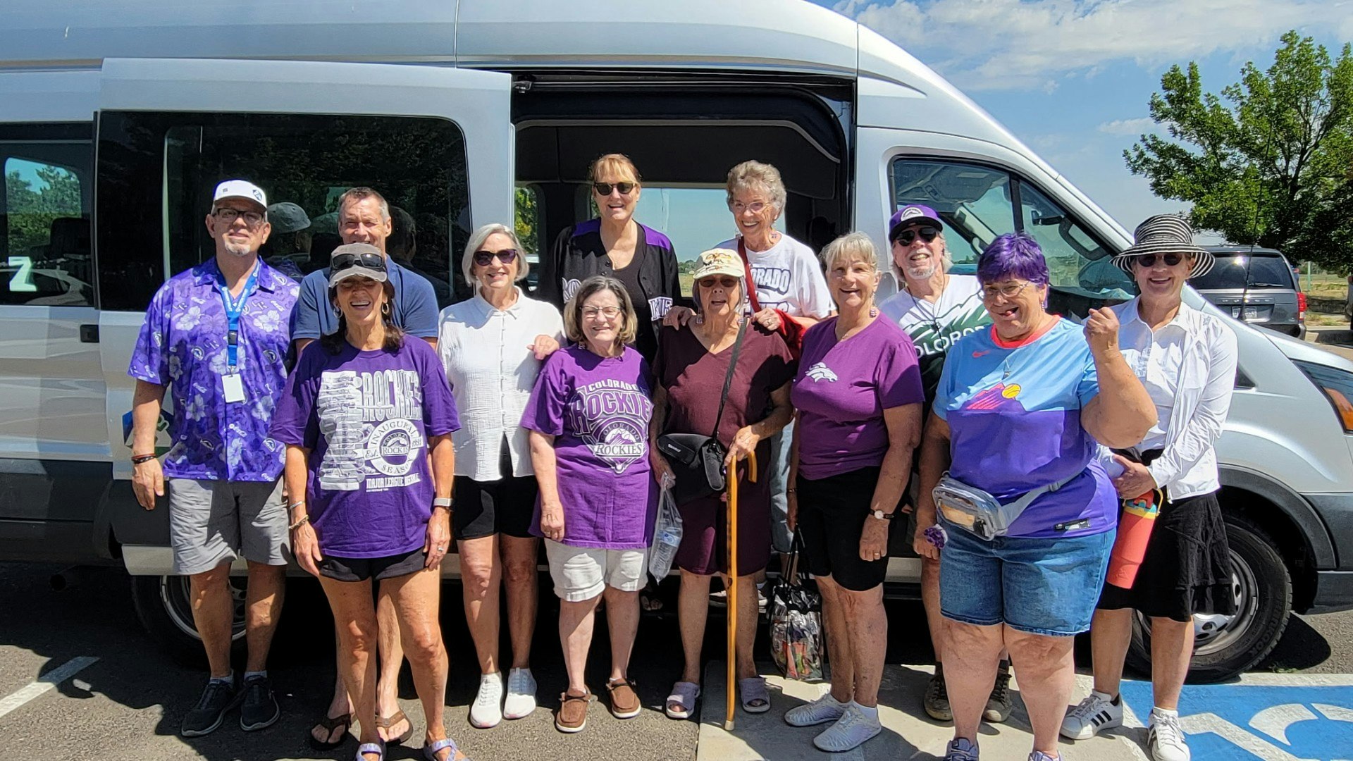 A group of people, mostly wearing purple shirts, poses together by a van on a sunny day.