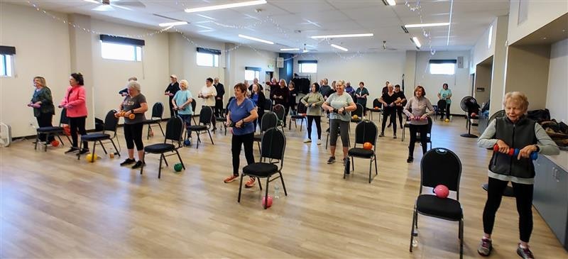 A group of people exercising in a spacious room, using colorful weights while seated or standing by chairs.