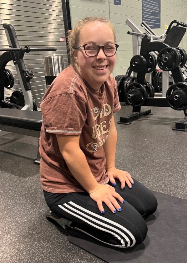 A person wearing glasses kneeling on a gym mat, smiling, with gym equipment in the background.