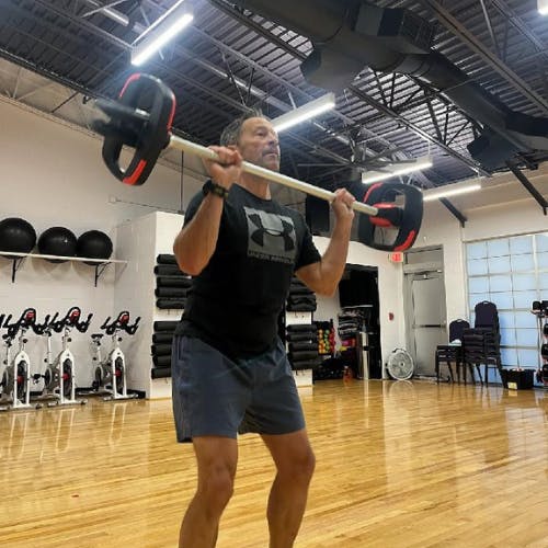 A person lifting weights in a gym, surrounded by exercise equipment and a wooden floor.