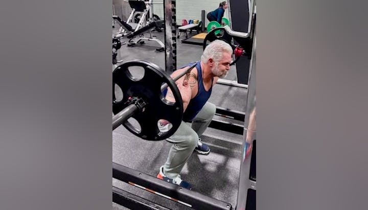 A man with a tattoo is doing a barbell squat in a gym setting.