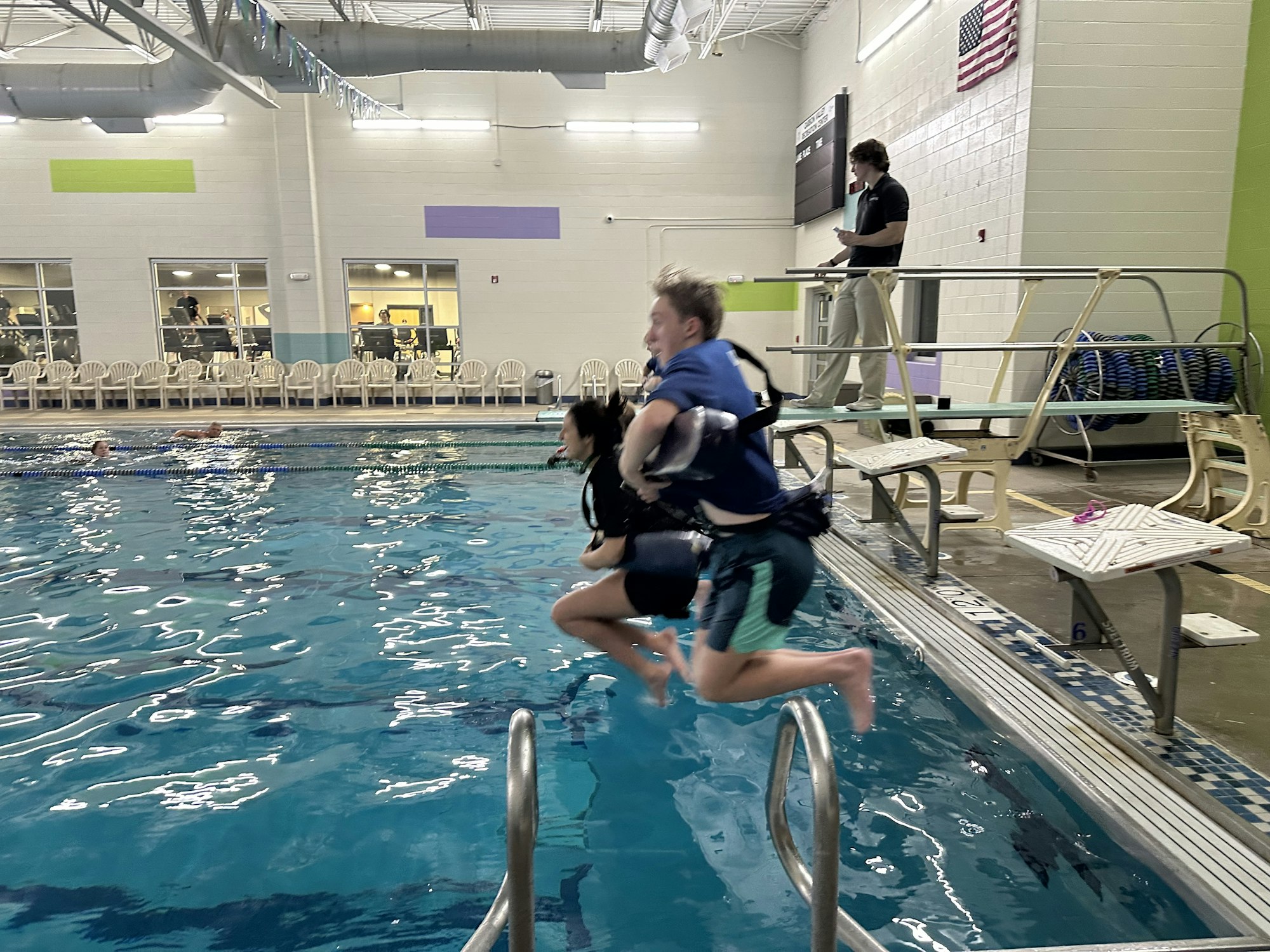 Two people jumping into an indoor swimming pool, with spectators in the background.