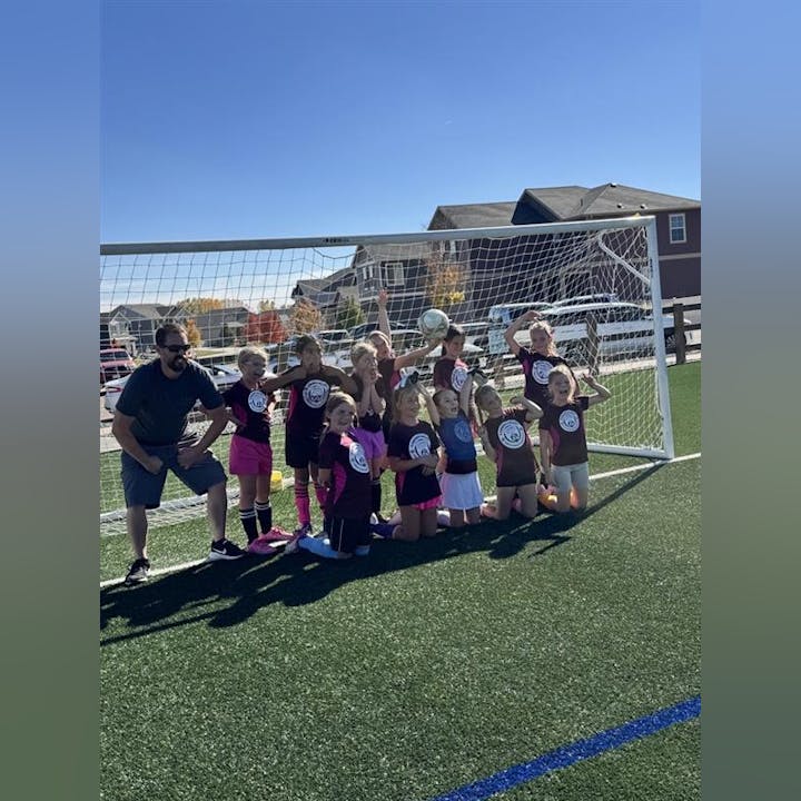 A group of young girls in soccer uniforms poses joyfully with a coach in front of a goal on a sunny field.