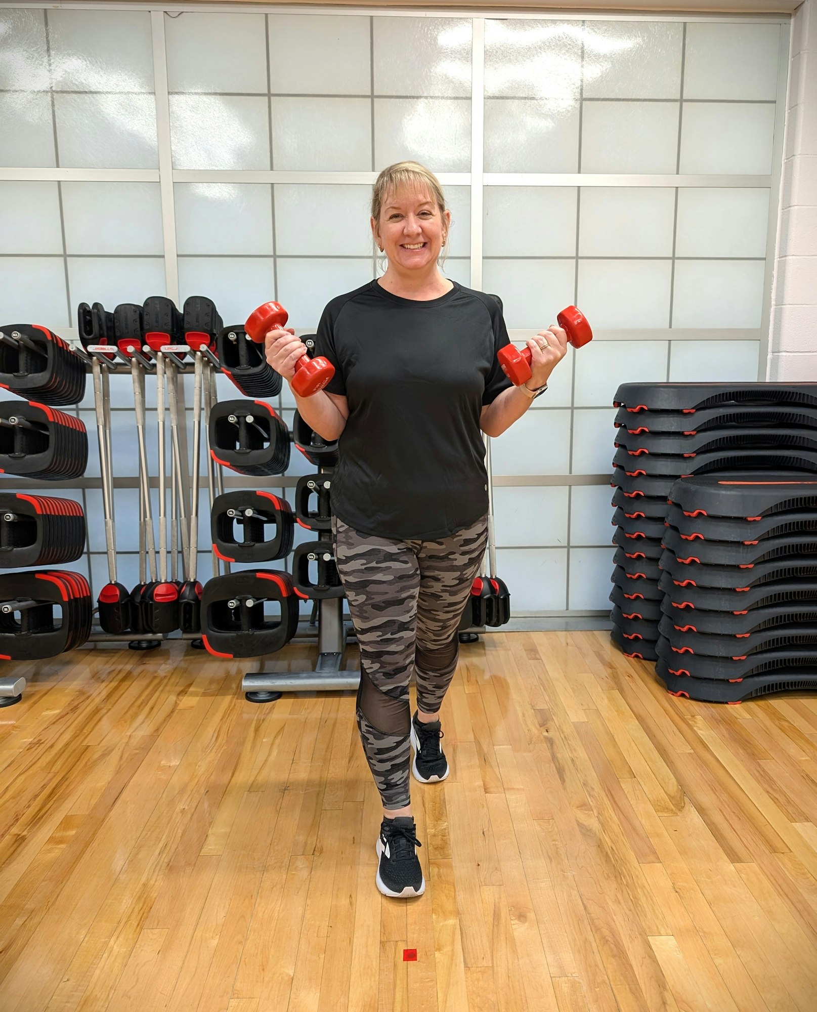A person holding red dumbbells in a gym, standing on a wooden floor with gym equipment in the background.