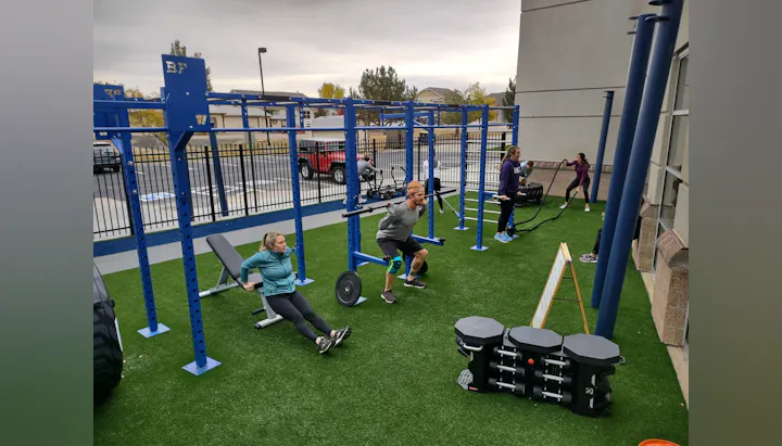 A group of people exercising outdoors, using various workout equipment like barbells and battle ropes in a gym setup.