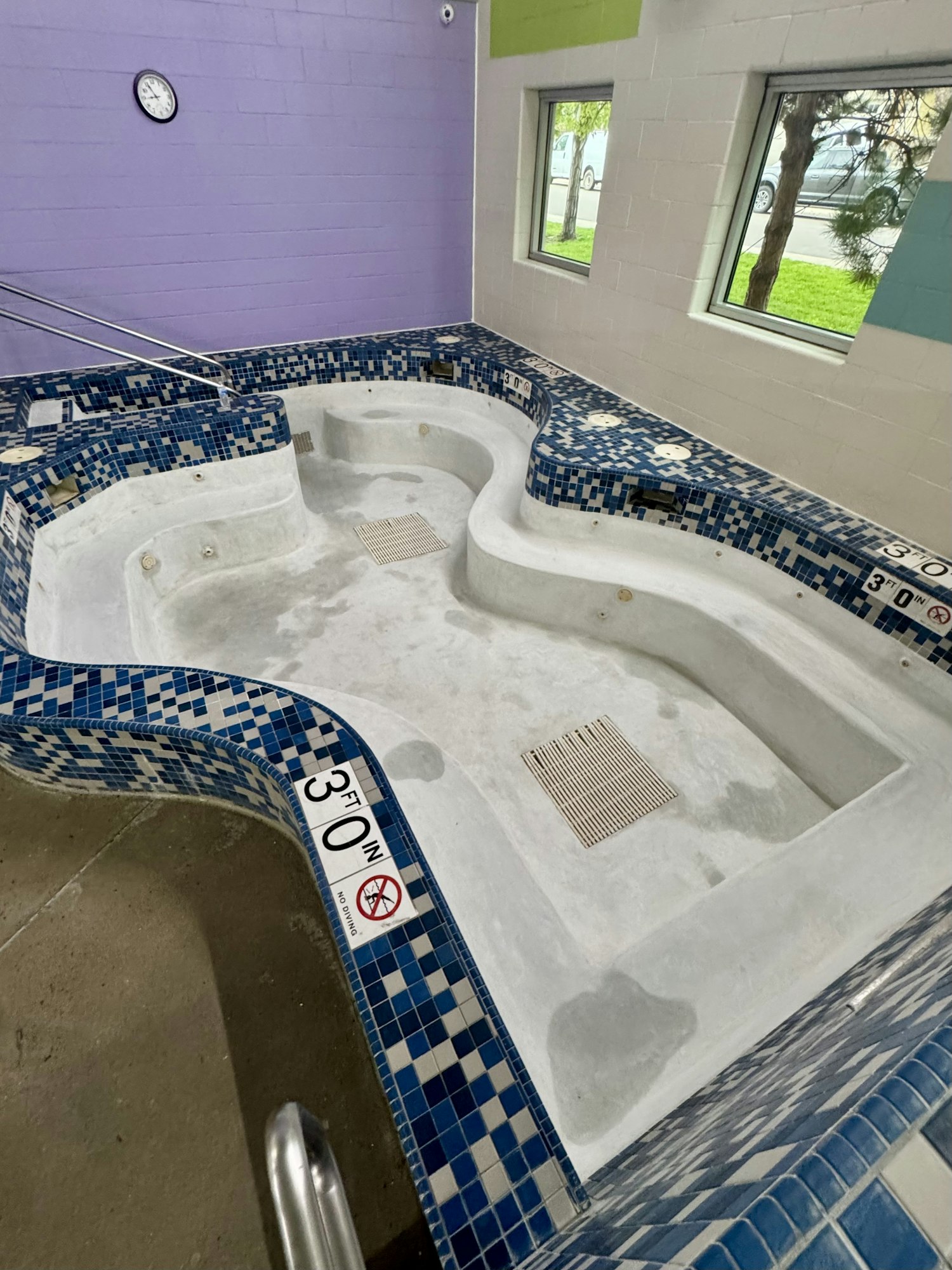 Empty tiled indoor hot tub, blue and white tiles, 3 ft deep sign, clock on wall, windows with outdoor view.