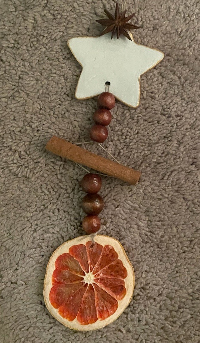A hanging decoration with a star, wooden beads, cinnamon stick, and a slice of dried orange, placed on a textured surface.