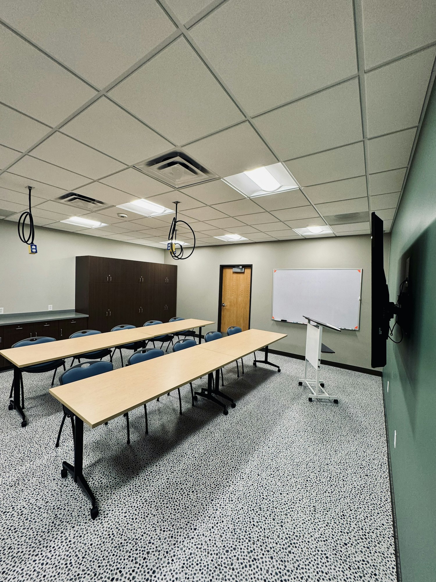 A classroom with two long tables, blue chairs, a podium, whiteboard, and ceiling projectors.