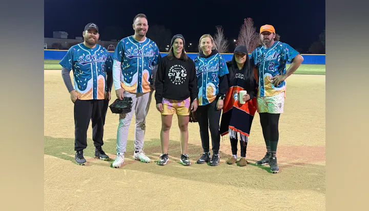 A group of six people in colorful, themed jerseys posing on a softball field at night.