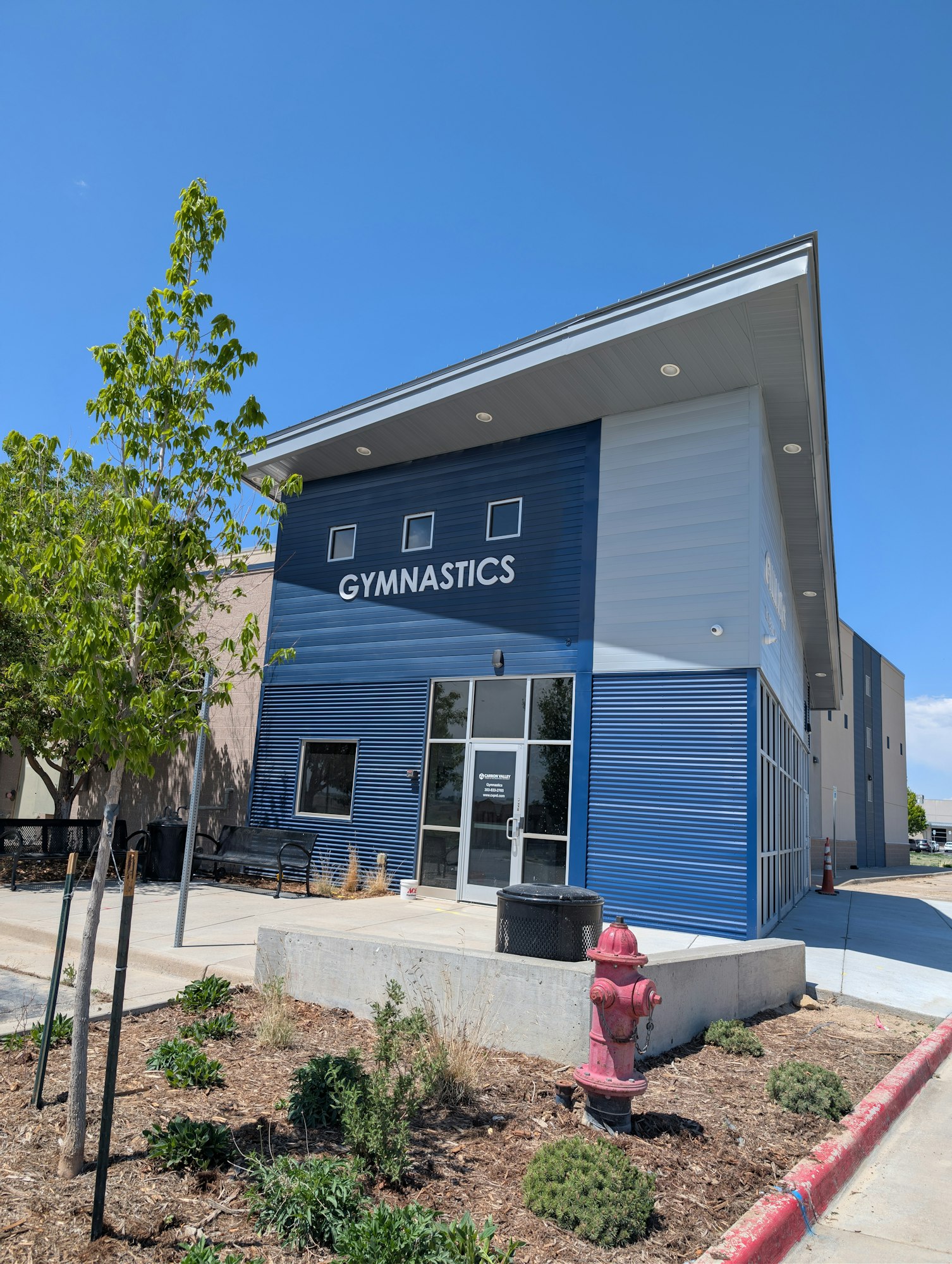 A modern gymnastics facility with blue siding, a welcoming entrance, and landscaping featuring a red fire hydrant.