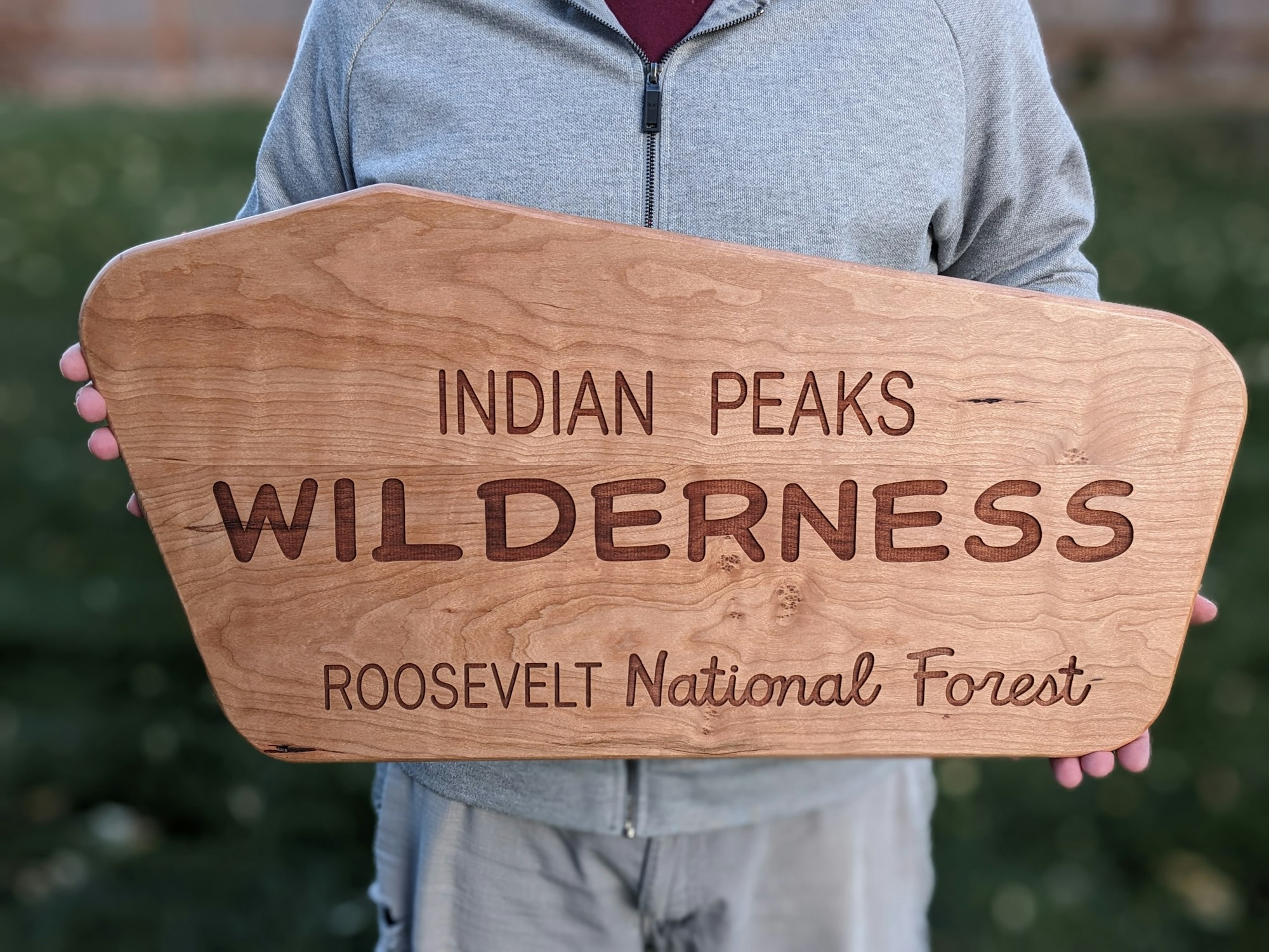 A person holds a wooden sign reading "Indian Peaks Wilderness, Roosevelt National Forest."