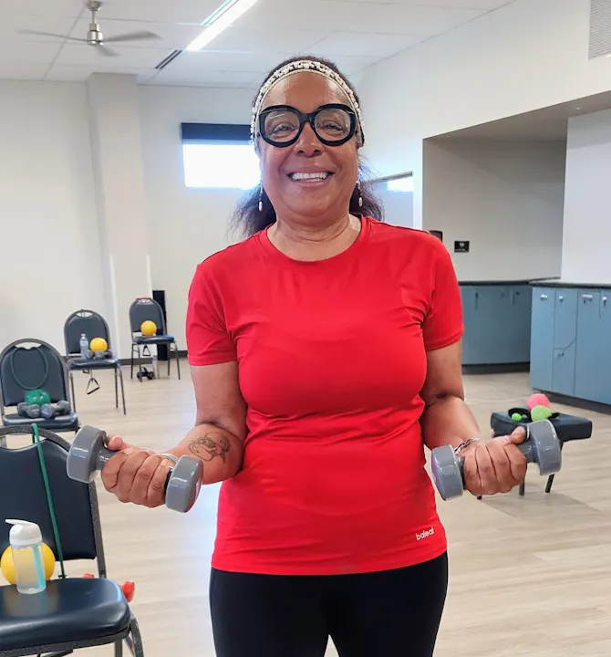 A smiling person in a red shirt holds dumbbells, standing in a fitness room with exercise equipment in the background.