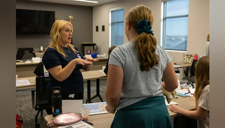 A woman is teaching a group, demonstrating something while others engage, possibly in a classroom or training setting.