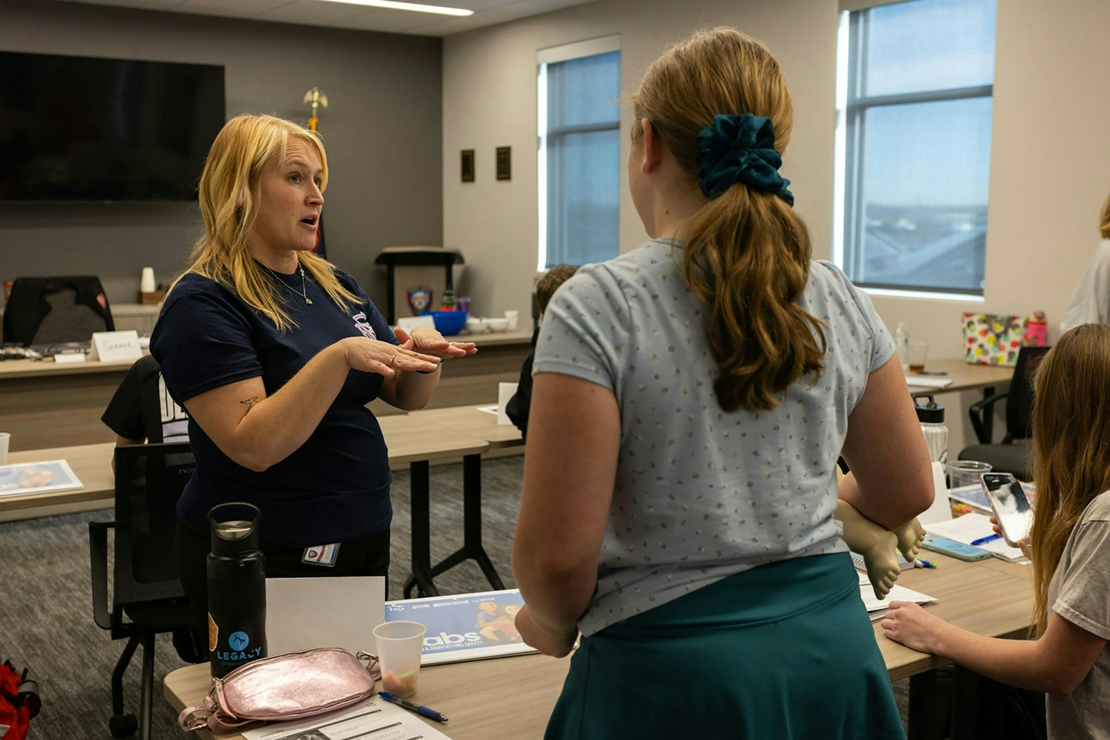 A woman is teaching a group, demonstrating something while others engage, possibly in a classroom or training setting.