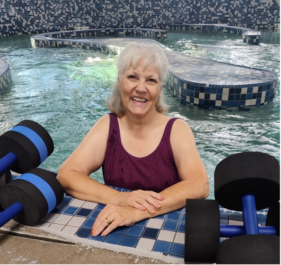A smiling woman in a burgundy tank top rests in an indoor pool with aquatic dumbbells.