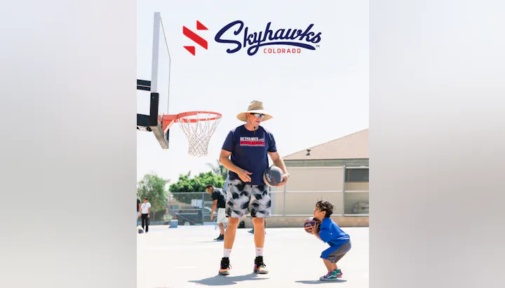 A coach teaches a young child basketball skills on an outdoor court, with others playing in the background.