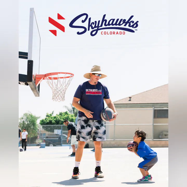 A coach teaches a young child basketball skills on an outdoor court, with others playing in the background.