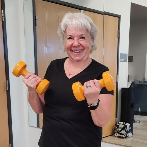 A smiling person holding two orange dumbbells in a room with mirrored doors.