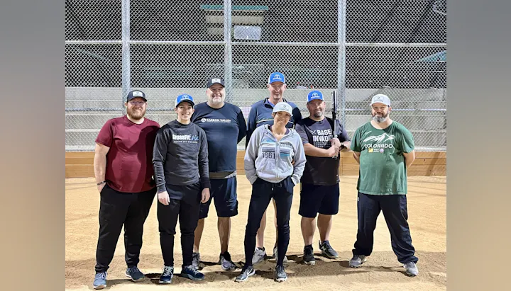 A group of seven people posing on a baseball field, wearing casual clothes and caps, with one holding a bat.
