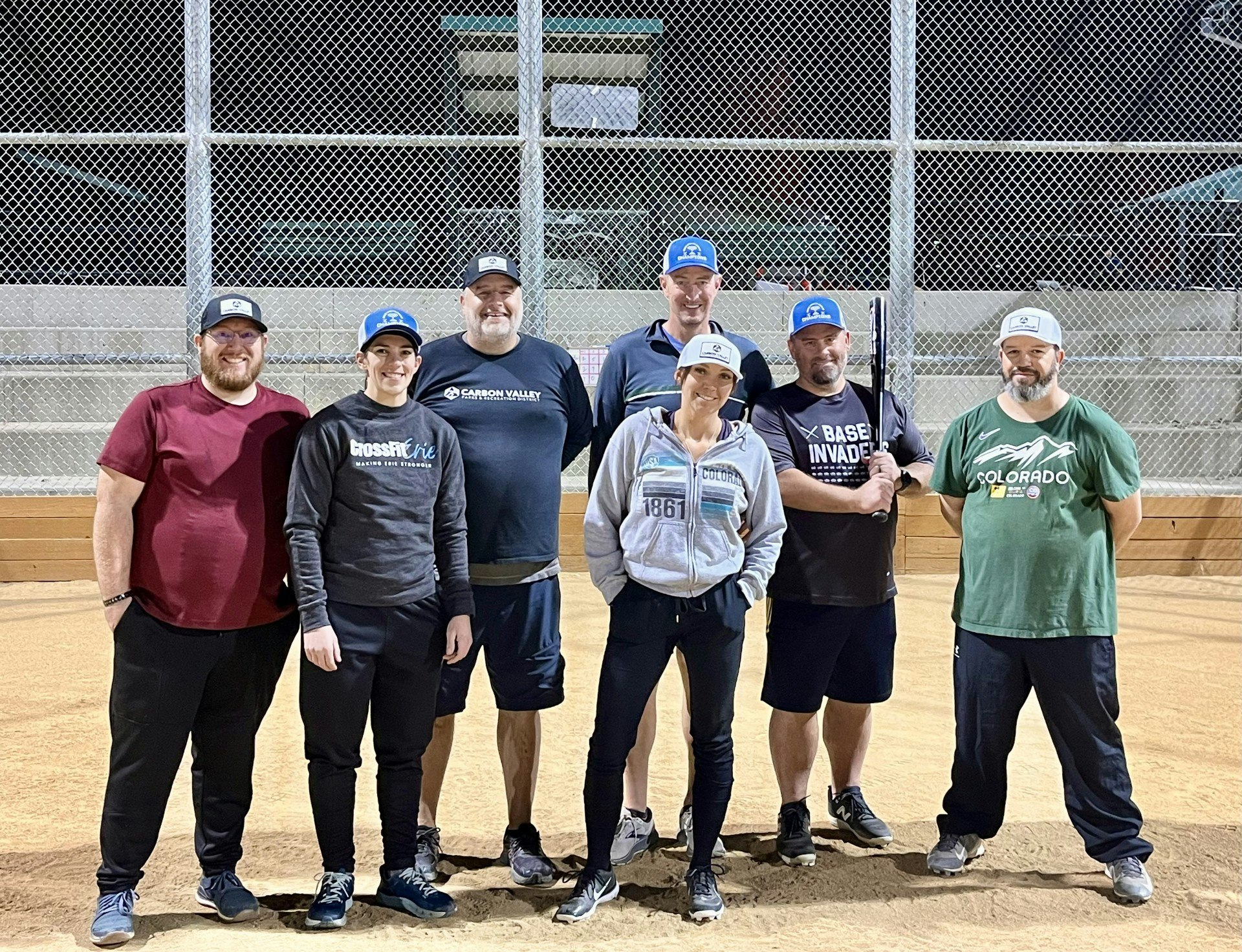 A group of seven people posing on a baseball field, wearing casual clothes and caps, with one holding a bat.