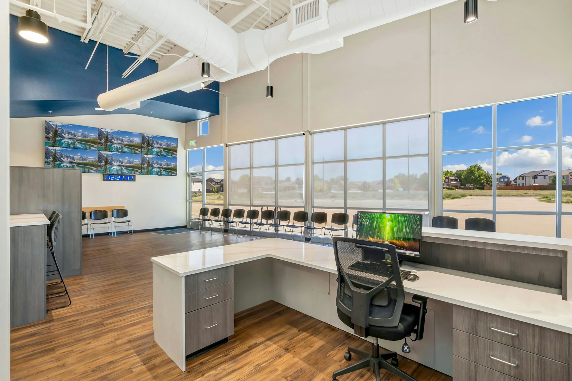 A modern office space featuring large windows, a workstation with a computer, multiple screens displaying images, and seating.